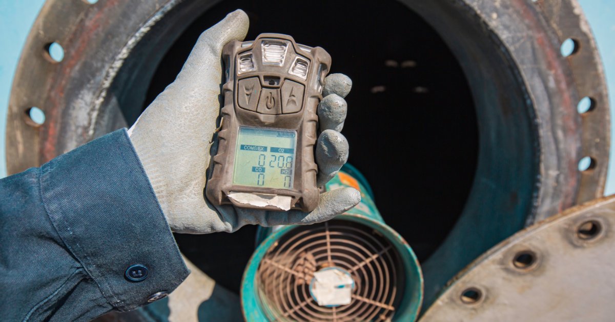 A worker wearing gray gloves holding a black handheld gas detector next to an open manhole on a wall.