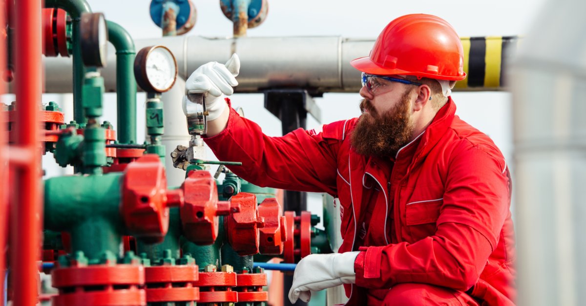 An employee wearing a red uniform and hard hat inspects a small monitoring device in an oil refinery.