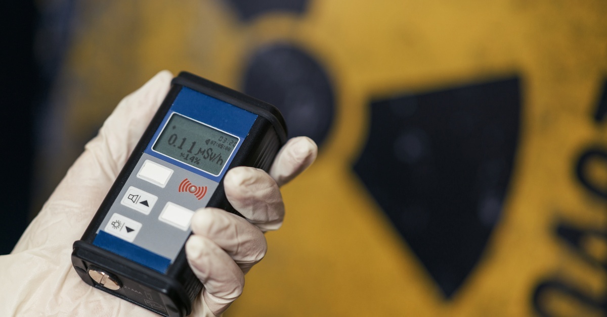 A gloved hand holds a digital dosimeter reading 0.11 mSv/h, with a yellow radiation hazard sign blurred in the background.