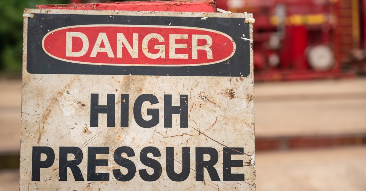 A weathered "Danger High Pressure" warning sign with red and black text on a worn white-and-black background, mounted outdoors.