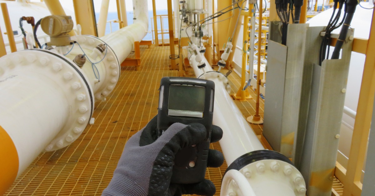 A worker uses a handheld gas monitor to check for hazardous gas levels along industrial piping on an offshore platform.