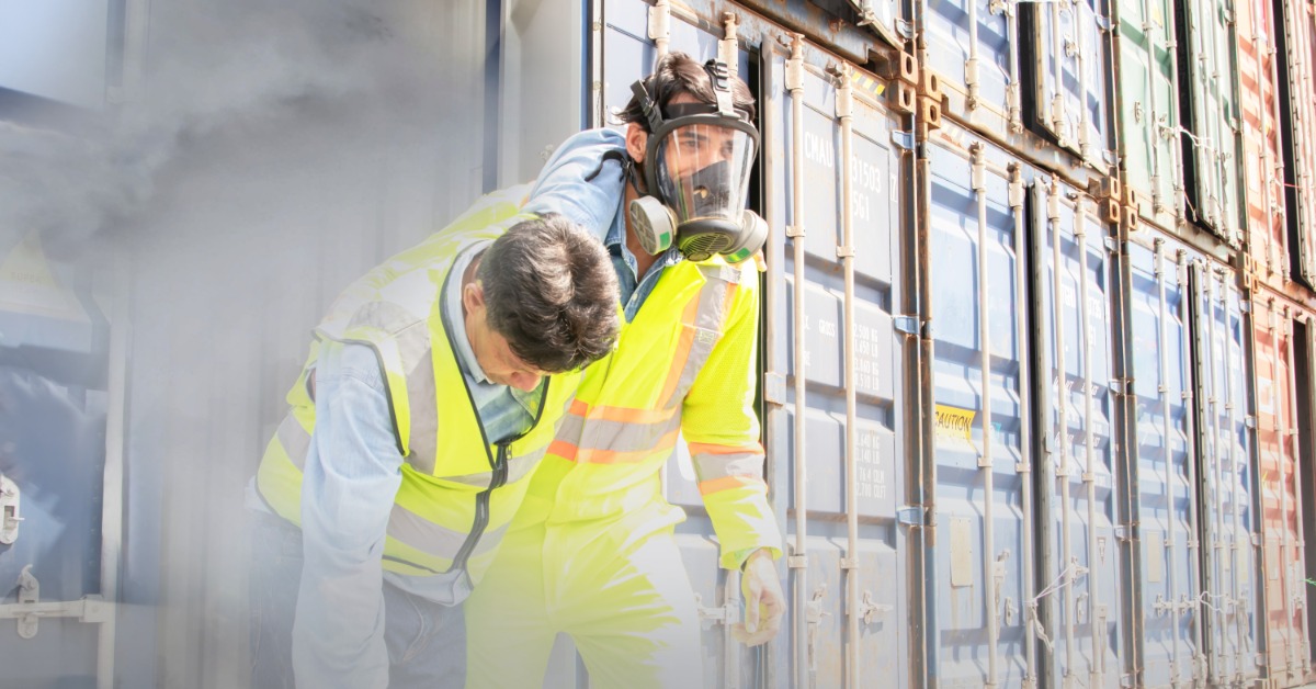 A rescue worker wearing a full-face gas mask assists a colleague through a smoky industrial hazard near cargo containers.
