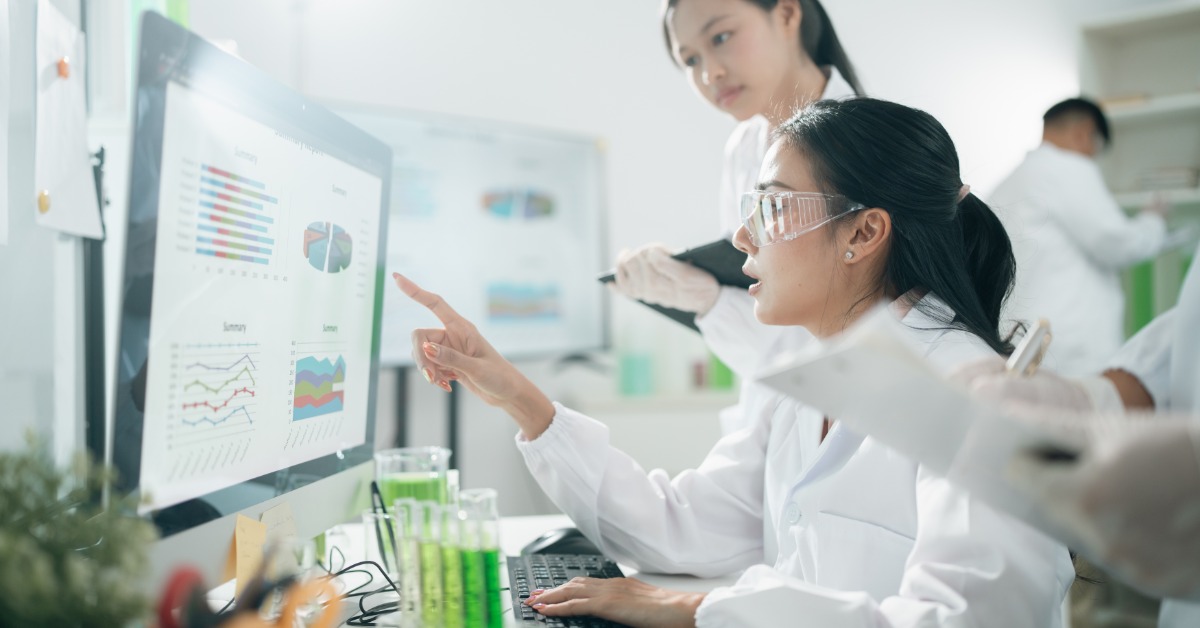 A Guide To Dose Rates and Radiation Exposure Limits Two scientists in white lab coats reviewing radiation data charts on a computer monitor with green test tubes in the foreground.