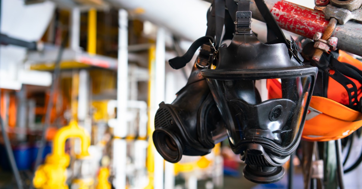 A Guide To Dose Rates and Radiation Exposure Limits Full-face radiation mask suspended from overhead metal piping in a plant facility with blurred yellow valves in the background.