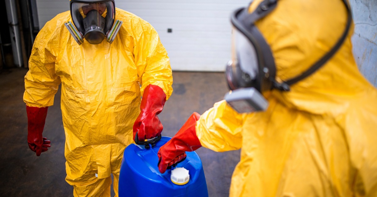 Two people in yellow protective suits and respirator masks handling a blue container in an industrial room.